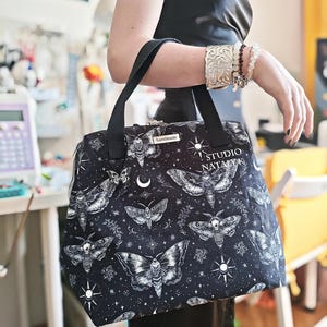 May include: A black tote bag featuring a white moth and celestial print with stars and crescent moons. The bag has black fabric handles, a silver zipper, and a small rectangular tag that reads handmade. The text STUDIO NATALY K is printed in white.