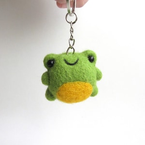 Peut inclure: Un porte-clés en forme de grenouille en feutre vert fait à la main, avec un ventre jaune. La grenouille a des yeux noirs et une bouche souriante. Le porte-clés est attaché à un anneau en métal argenté.