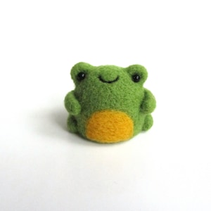 Könnte beinhalten: Ein kleiner grüner Filz-Frosch mit gelbem Bauch und schwarzen Augen. Der Frosch sitzt auf einem weißen Hintergrund.