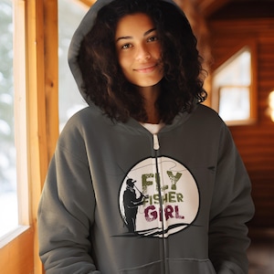 May include: A gray zip-up hoodie featuring a circular graphic with the words "FLY FISHER GIRL" and a silhouette of a person fly fishing. The hoodie has a hood and a white zipper.
