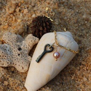 Peut inclure: Un collier en chaîne dorée avec un pendentif en forme de clé noire et une perle rose. Le collier repose sur une coquille blanche.