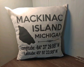 May include: White throw pillow with black text and an outline of Mackinac Island, Michigan. The text reads "Mackinac Island, Michigan" and includes the longitude and latitude coordinates: "Longitude: -84° 37' 29.99" W Latitude: 45° 51' 23.99" N"