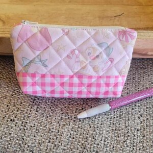 May include: A small, quilted pink pouch with a zipper closure. The pouch features a diamond pattern with cartoon images and a pink and white gingham bottom. A pink glitter pen rests beside it.