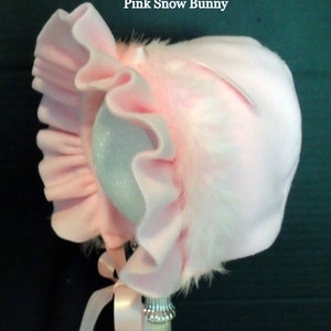 May include: Pink baby bonnet with a white fur trim and a ruffled edge. The bonnet is tied with a pink ribbon.
