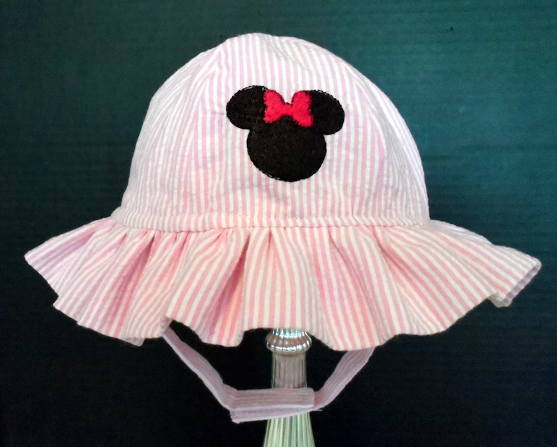 Op de afbeelding: Roze en witte gestreepte zonnehoed met een zwarte en rode Minnie Mouse applicatie.