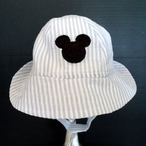 Op de afbeelding: Een blauwe en witte gestreepte zonnehoed met een zwart Mickey Mouse silhouet aan de voorkant.