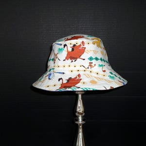 May include: A white bucket hat featuring a repeating pattern of cartoon characters including a brown warthog, a small meerkat, and various tropical plants. The design includes geometric diamond shapes and small sun icons in orange and teal colors.