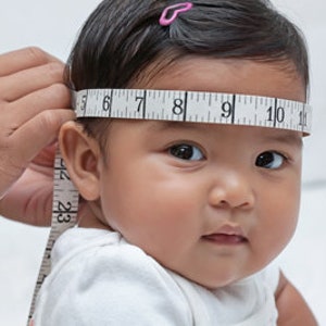 Op de afbeelding: Het hoofd van een baby wordt gemeten met een meetlint. Het meetlint is om het hoofd van de baby gewikkeld en de nummers 6, 7, 8, 9, 10 en 11 zijn zichtbaar.