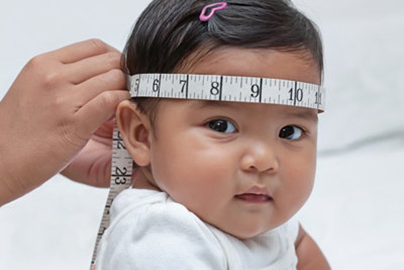 Op de afbeelding: Het hoofd van een baby wordt gemeten met een meetlint. Het meetlint is om het hoofd van de baby gewikkeld en de nummers 6, 7, 8, 9, 10 en 11 zijn zichtbaar.