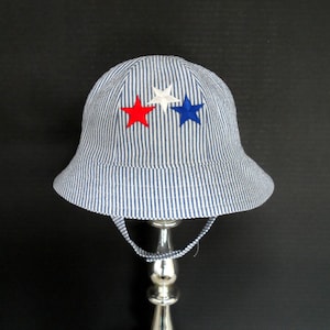 May include: A blue and white striped bucket hat with three embroidered stars in red, white, and blue. The hat has a chin strap and a wide brim. The hat is displayed on a silver stand.