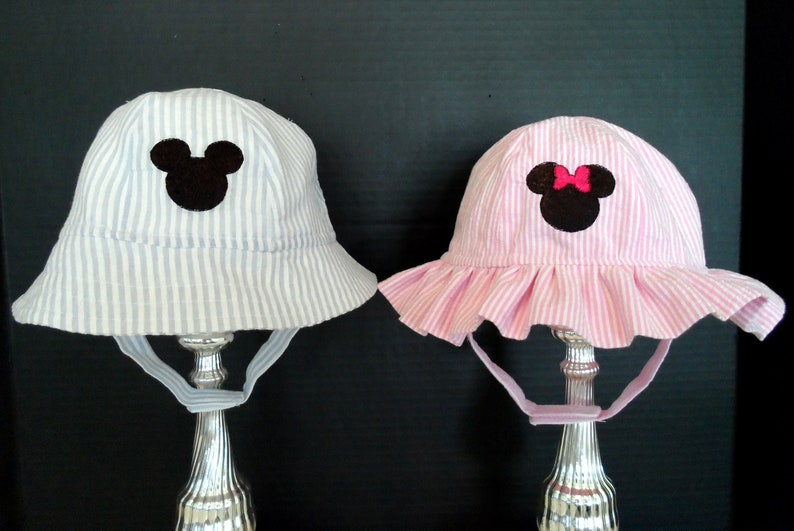 Op de afbeelding: Twee gestreepte zonnehoeden met Mickey Mouse en Minnie Mouse applicaties. De Mickey Mouse hoed is blauw-wit gestreept en de Minnie Mouse hoed is roze-wit gestreept.