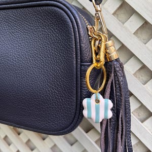 Puede incluir: Un bolso bandolera de cuero granulado azul marino oscuro con un cierre de metal dorado y una borla de cuero a juego. Un pequeño dije en forma de flor con rayas verticales blancas y azul claro cuelga del anillo dorado.