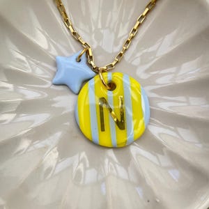 Puede incluir: Un collar de cadena dorada con un colgante redondo de rayas verticales amarillas y azul claro, con una letra N dorada en el centro. Un pequeño dije en forma de estrella azul claro cuelga de la misma anilla dorada que el colgante redondo.