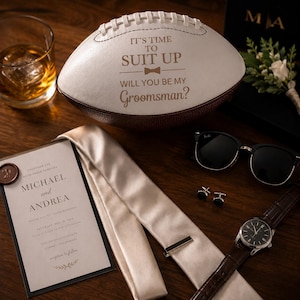 Puede incluir: Un bal&oacute;n de f&uacute;tbol americano blanco y marr&oacute;n grabado con el texto IT'S TIME TO SUIT UP WILL YOU BE MY Groomsman? junto a una invitaci&oacute;n de boda para Michael y Andrea, una corbata color champ&aacute;n, gafas de sol negras, gemelos y un reloj.