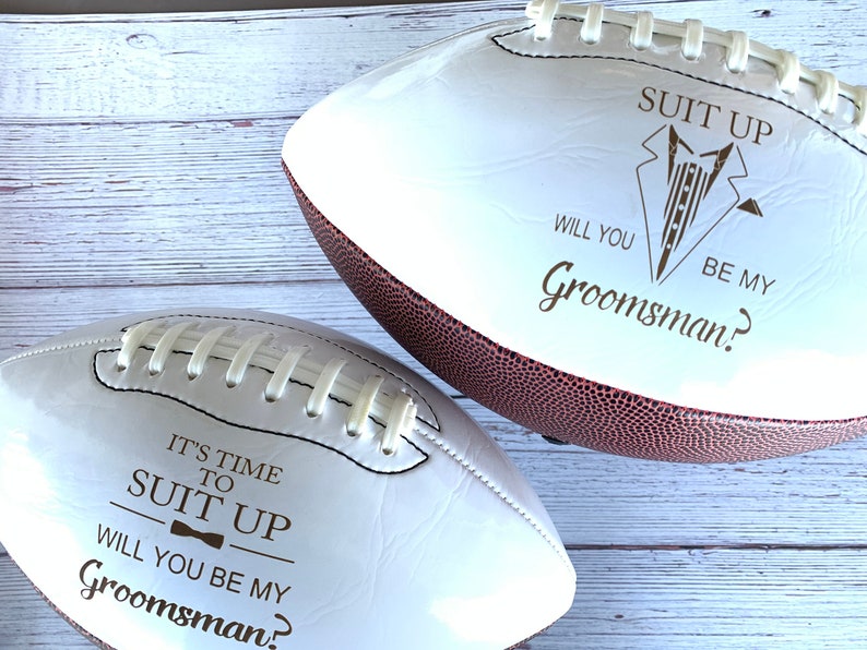 May include: Two white footballs with brown stitching. One football has the text "It's time to suit up. Will you be my groomsman?" The other football has the text "Suit up. Will you be my groomsman?"
