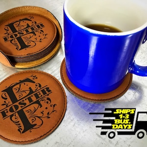 May include: Set of four brown leather coasters with a black floral design and the name "Foster" in a script font. The coasters are stacked on top of each other and are sitting on a white surface. A blue mug with coffee is sitting on a brown leather coaster.