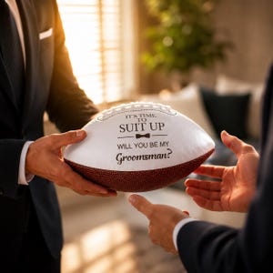 Puede incluir: Un bal&oacute;n de f&uacute;tbol americano blanco y marr&oacute;n con texto negro que dice IT'S TIME TO SUIT UP, un peque&ntilde;o icono de pajarita y la pregunta WILL YOU BE MY Groomsman? El bal&oacute;n tiene cordones blancos y una superficie de cuero marr&oacute;n texturizado.