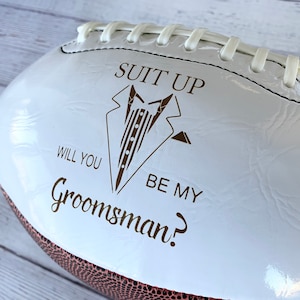 Puede incluir: Un bal&oacute;n de f&uacute;tbol americano blanco brillante con un gr&aacute;fico marr&oacute;n de una chaqueta de esmoquin y el texto "SUIT UP WILL YOU BE MY Groomsman?". El bal&oacute;n tiene cordones blancos en la costura superior y una secci&oacute;n de cuero marr&oacute;n texturizado visible en el borde inferior.