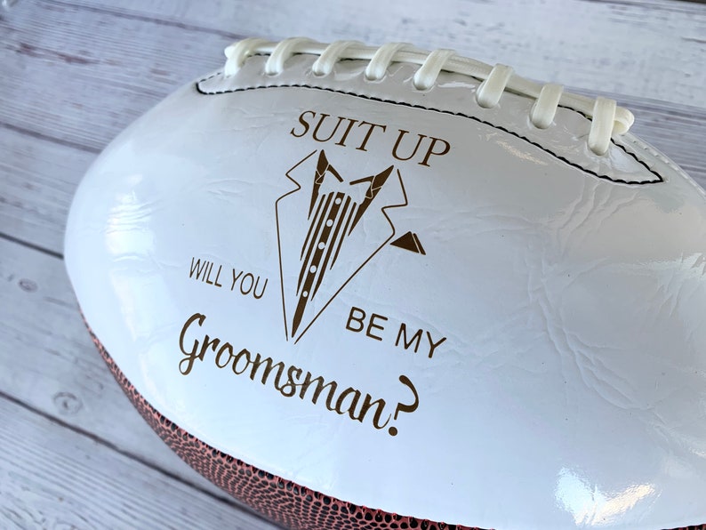 Puede incluir: Un bal&oacute;n de f&uacute;tbol americano blanco brillante con un gr&aacute;fico marr&oacute;n de una chaqueta de esmoquin y el texto "SUIT UP WILL YOU BE MY Groomsman?". El bal&oacute;n tiene cordones blancos en la costura superior y una secci&oacute;n de cuero marr&oacute;n texturizado visible en el borde inferior.