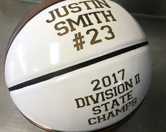 May include: White basketball with black lines. The basketball has gold lettering that reads "Justin Smith #23 2017 Division II State Champs".