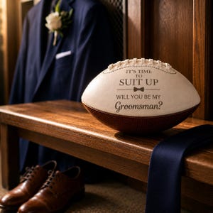 Puede incluir: Un bal&oacute;n de f&uacute;tbol americano blanco y marr&oacute;n con texto negro que dice IT'S TIME TO SUIT UP WILL YOU BE MY Groomsman? junto a un icono de pajarita. El bal&oacute;n presenta cordones blancos y un panel de cuero marr&oacute;n texturizado en la base.