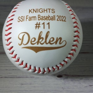 Op de afbeelding: Witte baseball met rode stiksels. De bal is gepersonaliseerd met gouden letters die "Knights SSI Farm Baseball 2022 #11 Deklen" lezen.