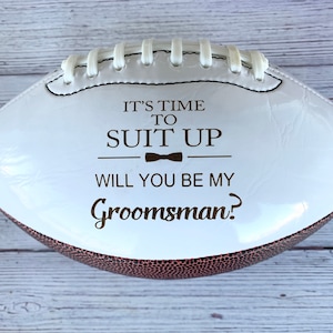 Puede incluir: Un bal&oacute;n de f&uacute;tbol americano blanco y marr&oacute;n con cordones blancos y texto marr&oacute;n oscuro que dice IT'S TIME TO SUIT UP, un peque&ntilde;o icono de pajarita y la pregunta WILL YOU BE MY Groomsman? La superficie del bal&oacute;n es lisa y brillante.