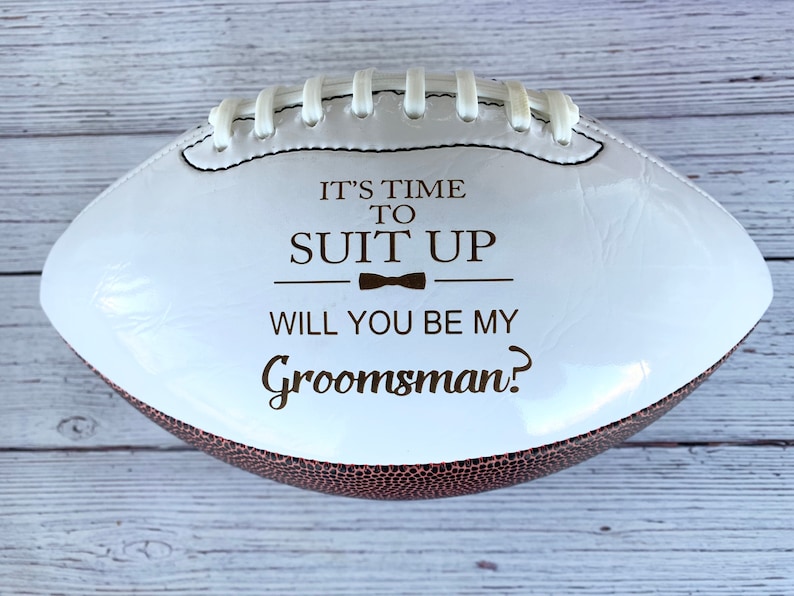 Puede incluir: Un bal&oacute;n de f&uacute;tbol americano blanco y marr&oacute;n con cordones blancos y texto marr&oacute;n oscuro que dice IT'S TIME TO SUIT UP, un peque&ntilde;o icono de pajarita y la pregunta WILL YOU BE MY Groomsman? La superficie del bal&oacute;n es lisa y brillante.