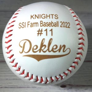 Peut inclure: Balle de baseball blanche avec des coutures rouges. La balle est personnalisée avec du texte doré qui dit "Knights SSI Farm Baseball 2022 #11 Deklen".