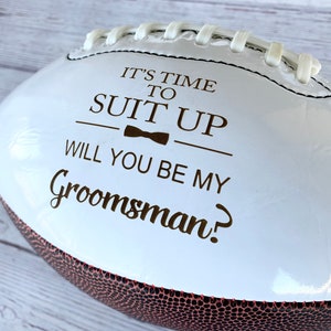 Puede incluir: Un bal&oacute;n de f&uacute;tbol americano blanco con paneles marrones texturizados y cordones blancos. La superficie presenta texto marr&oacute;n oscuro que dice IT'S TIME TO SUIT UP, un peque&ntilde;o icono de pajarita y el texto WILL YOU BE MY Groomsman? impreso.