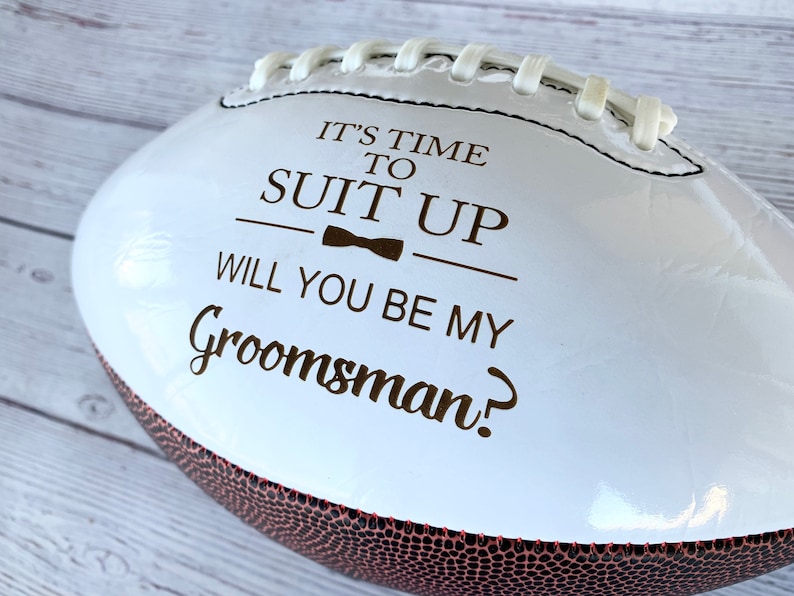 Puede incluir: Un bal&oacute;n de f&uacute;tbol americano blanco con paneles marrones texturizados y cordones blancos. La superficie presenta texto marr&oacute;n oscuro que dice IT'S TIME TO SUIT UP, un peque&ntilde;o icono de pajarita y el texto WILL YOU BE MY Groomsman? impreso.