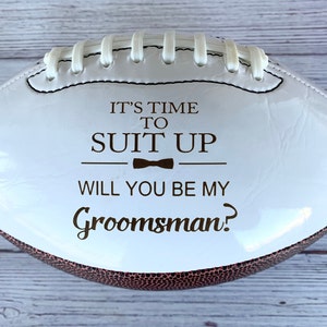 Puede incluir: Un bal&oacute;n de f&uacute;tbol americano blanco con extremos texturizados en marr&oacute;n y cordones blancos. El centro presenta el texto en marr&oacute;n oscuro IT'S TIME TO SUIT UP, un icono de pajarita y el texto WILL YOU BE MY Groomsman? en una combinaci&oacute;n de fuentes.