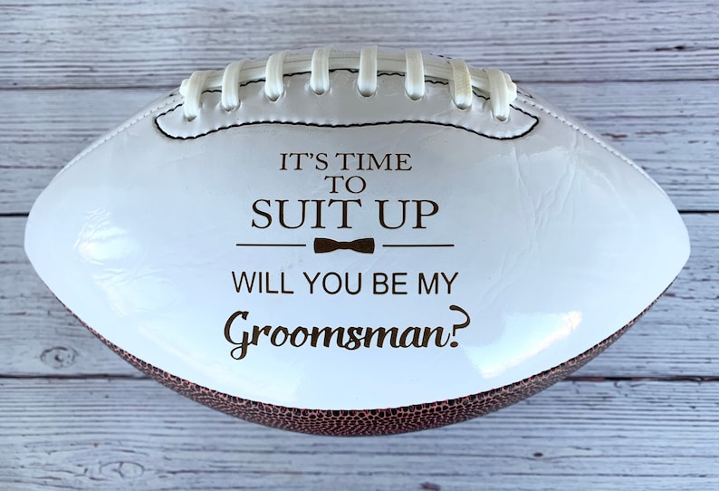 Puede incluir: Un bal&oacute;n de f&uacute;tbol americano blanco con extremos texturizados en marr&oacute;n y cordones blancos. El centro presenta el texto en marr&oacute;n oscuro IT'S TIME TO SUIT UP, un icono de pajarita y el texto WILL YOU BE MY Groomsman? en una combinaci&oacute;n de fuentes.
