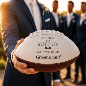 May include: A white and brown leather American football featuring black printed text that reads IT'S TIME TO SUIT UP WILL YOU BE MY Groomsman? with a small bow tie graphic centred between the lines of text on the surface of the ball.