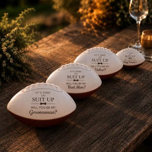 Puede incluir: Tres balones de f&uacute;tbol americano blancos y marrones con texto negro que dice IT'S TIME TO SUIT UP seguido de WILL YOU BE MY GROOMSMAN, WILL YOU BE MY BEST MAN o WILL YOU BE MY USHER, adem&aacute;s de un bal&oacute;n m&aacute;s peque&ntilde;o con WILL YOU BE MY RING BEARER.
