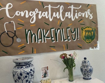 May include: A brown banner with white lettering that reads "Congratulations Makinley!" with a green and gold bear illustration. A stethoscope is drawn on the left side of the banner. The banner is hanging on a white wall. A white table with a blue and white porcelain vase with flowers, a blue and white porcelain jar, and a gold "GRAD" sign.
