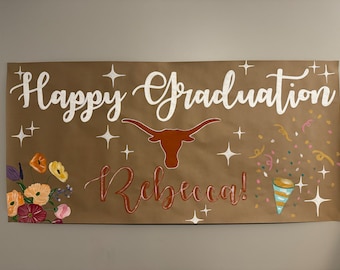 May include: A brown paper banner with the text "Happy Graduation Rebecca!" in white cursive lettering. The banner also features a brown and orange Longhorn steer head, white stars, and colorful confetti.