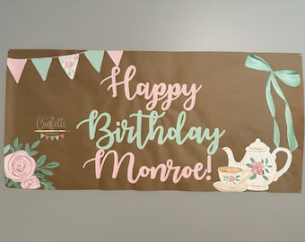 May include: A brown banner with the text "Happy Birthday Monroe!" in pink and green lettering. The banner also features a floral design with pink and green flowers, a teacup and teapot with floral designs, and a green ribbon bow.