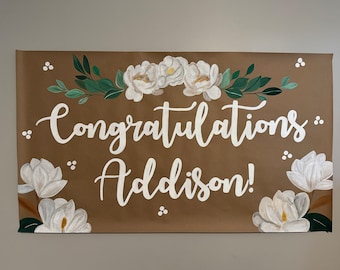 May include: A brown paper banner with white lettering that reads "Congratulations Addison!"  The banner is decorated with white flowers and green leaves.