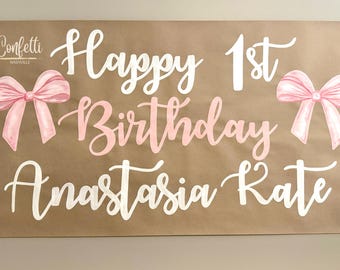 May include: A brown banner with the text "Happy 1st Birthday Anastasia Kate" in white and pink script. Two pink bows and floral accents frame the text. The banner is for a birthday celebration.