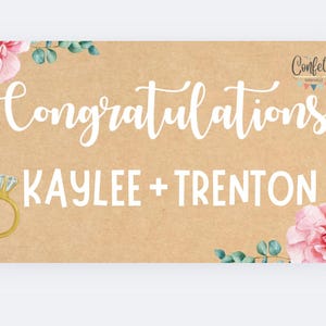 Puede incluir: Un letrero rectangular con fondo de papel kraft. El letrero presenta la palabra "Enhorabuena" en escritura blanca, junto con los nombres "KAYLEE + TRENTON" en negrita. Una ilustración de anillo dorado está a la izquierda, y acentos florales enmarcan la parte superior e inferior.