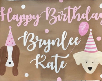 May include: A brown paper banner with the text "Happy Birthday Brynlee Kate" in pink and white lettering. The banner features two dog illustrations wearing pink and white party hats. One dog is brown and white, and the other is white with a pink balloon.