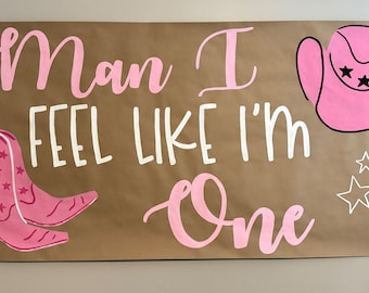 Può includere: Uno striscione di carta marrone con il testo "Man I feel like I'm one" scritto in vernice rosa e bianca. Ci sono stivali da cowboy rosa e un cappello da cowboy rosa con stelle bianche.