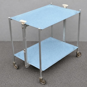 Może przedstawiać: Dwupoziomowy wózek do serwowania w stylu vintage z jasnoniebieskimi, teksturowanymi półkami. Wózek ma srebrną metalową ramę i małe kółka. Białe gałki znajdują się na górnych rogach. Ma około 60 cm długości.