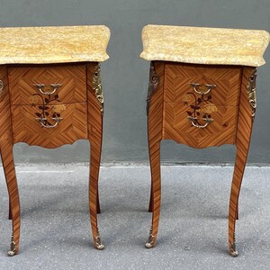 May include: A pair of antique bedside tables with a light-colored marble top. The wooden cabinets feature a geometric pattern, floral inlay, and ornate brass hardware. The legs are curved and end in brass feet.