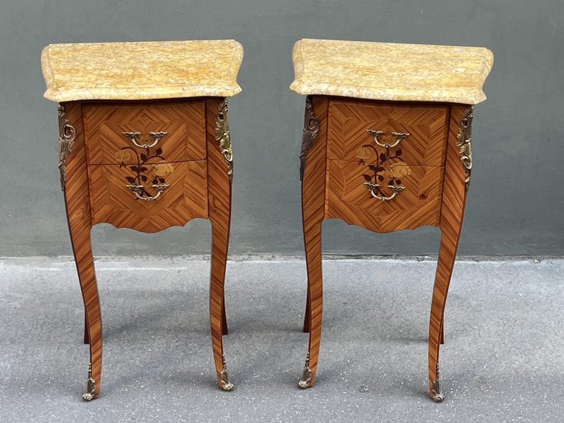 May include: A pair of antique bedside tables with a light-colored marble top. The wooden cabinets feature a geometric pattern, floral inlay, and ornate brass hardware. The legs are curved and end in brass feet.