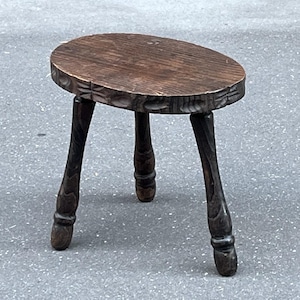 May include: A small, antique wooden stool with a dark brown finish. The oval seat has a carved edge, and the three legs are curved and detailed. This decorative stool is a vintage piece.