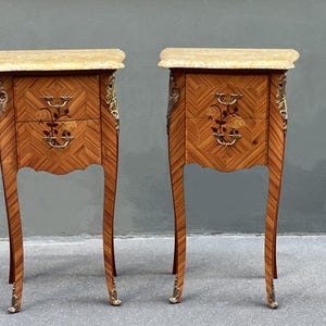 May include: Pair of antique wooden nightstands with marble tops. Each nightstand features two drawers with floral inlay and ornate brass hardware. The legs are curved and end in brass feet. The wood has a herringbone pattern.
