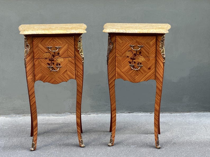 May include: Pair of antique wooden nightstands with marble tops. Each nightstand features two drawers with floral inlay and ornate brass hardware. The legs are curved and end in brass feet. The wood has a herringbone pattern.