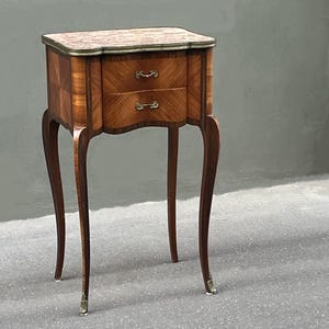 Può includere: Comodino antico in legno con due cassetti. Ha un piano in marmo e ferramenta in ottone. Le gambe curve terminano con piedini in ottone. Il legno ha un caldo tono marrone-rossastro. Il tavolo è alto circa 60 cm.